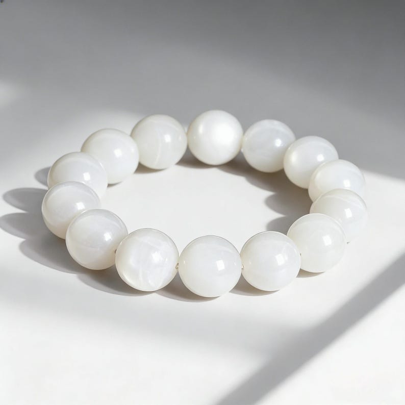 May include: A white beaded bracelet with large, round beads. The bracelet is made of smooth, polished beads, and is displayed on a white surface. The beads have a slight sheen, reflecting the light.