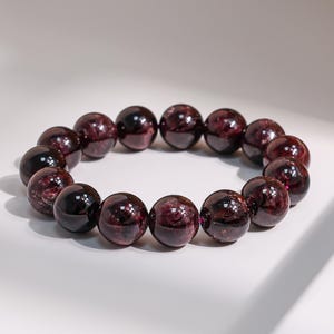 Peut inclure: Un bracelet composé de perles de grenat polies, de couleur rouge foncé. Les perles rondes sont enfilées ensemble, formant une forme circulaire. Les pierres brillantes sont exposées sur une surface blanche. Chaque perle mesure environ 1,5 cm de diamètre.