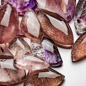 May include: A collection of teardrop-shaped, faceted, translucent gemstone pendants in shades of purple, brown, and pink. The stones have a unique, cloudy, and striated appearance.