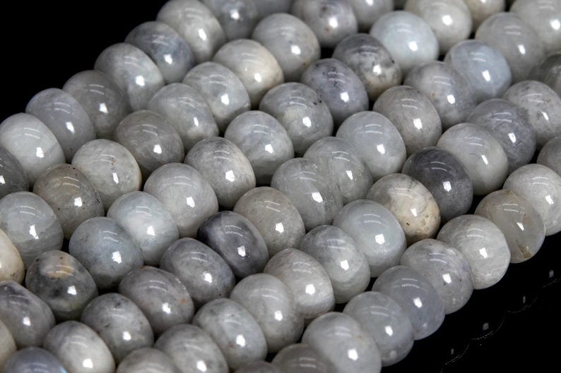 May include: Close-up of a strand of smooth, oval-shaped beads. The beads are a mix of pale gray and white, with a glossy finish. The beads are strung together, creating a pattern of light and shadow.