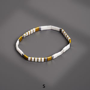 May include: A white and gold beaded bracelet with rectangular beads. The bracelet has a simple design with alternating white and gold-colored beads, and a few gray beads. The bracelet is shown on a gray background.