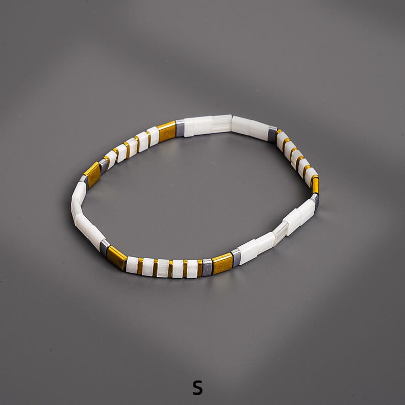 May include: A white and gold beaded bracelet with rectangular beads. The bracelet has a simple design with alternating white and gold-colored beads, and a few gray beads. The bracelet is shown on a gray background.