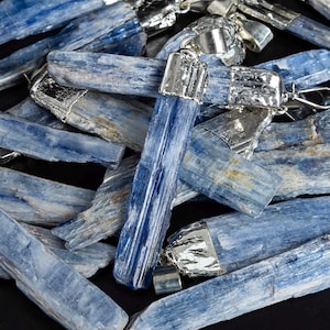Pode incluir: Um grupo de pingentes de cristal de cianita azul com tampas prateadas. Os cristais são longos e finos, com uma textura estriada. Os pingentes são perfeitos para fazer joias ou usar na cura com cristais.