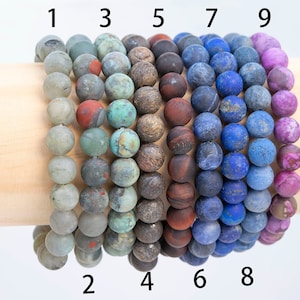 May include: Nine bracelets made of different types of stones. The bracelets are stacked on a wooden display stand. The bracelets are numbered 1 through 9.