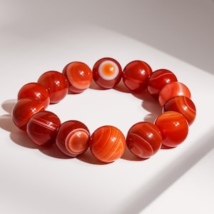 May include: A beaded bracelet featuring large, round beads in shades of red and orange. The beads have a polished, glossy finish and display unique patterns of stripes and swirls. The bracelet is displayed on a white surface.