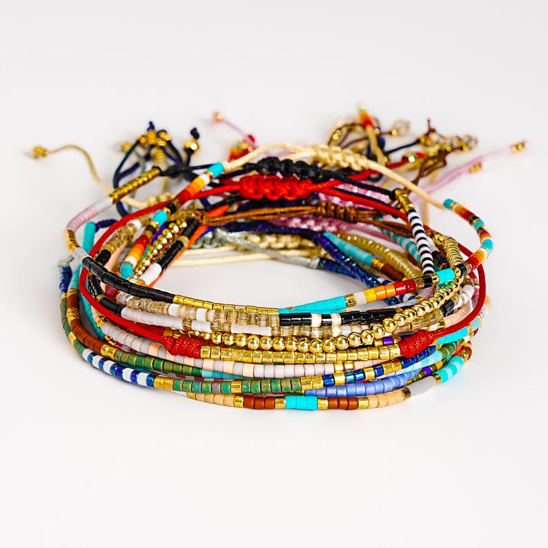 May include: A stack of colorful beaded bracelets. The bracelets feature a variety of colors including gold, red, blue, and turquoise. The beads are small and strung together on a cord. Some bracelets have adjustable knots.