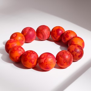 Könnte beinhalten: Ein kreisförmiges Armband aus elf lebendigen, marmorierten Perlen. Die Perlen sind hauptsächlich orange mit roten und kastanienbraunen Akzenten, wodurch ein einzigartiges, auffälliges Muster entsteht. Das Armband wird auf einer weißen Oberfläche präsentiert.