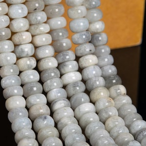 May include: Strands of light gray, barrel-shaped beads. The beads have a smooth, polished appearance and are strung together, creating a flowing, textured pattern. The beads are a neutral color, suitable for jewelry making.