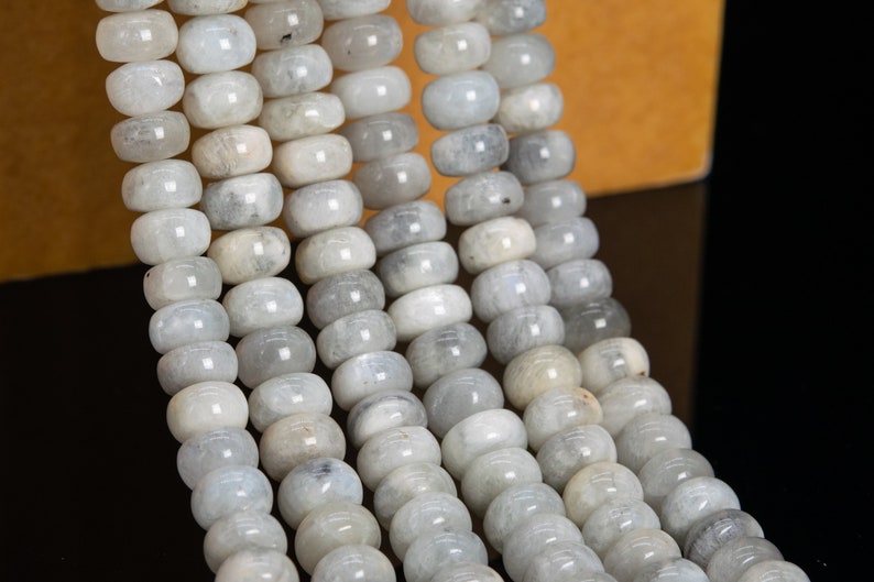 May include: Strands of light gray, barrel-shaped beads. The beads have a smooth, polished appearance and are strung together, creating a flowing, textured pattern. The beads are a neutral color, suitable for jewelry making.