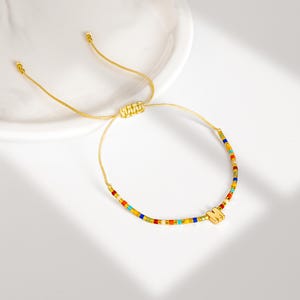 May include: A gold-colored adjustable bracelet with a beaded design. The beads are small and rectangular, in a variety of colors including red, yellow, blue, and green. A gold-colored charm in the shape of the letter "M" is attached to the bracelet.