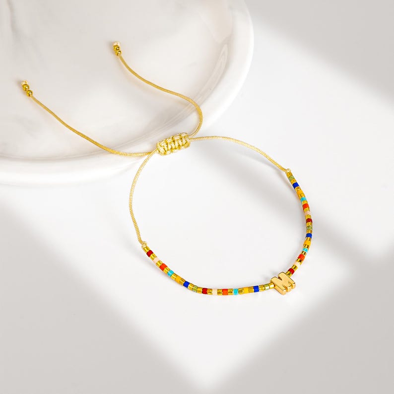 May include: A gold-colored adjustable bracelet with a beaded design. The beads are small and rectangular, in a variety of colors including red, yellow, blue, and green. A gold-colored charm in the shape of the letter "M" is attached to the bracelet.
