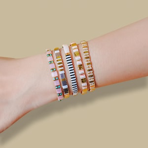 May include: A collection of six colorful bracelets adorns a wrist. The bracelets feature a variety of designs, including stripes, solid colors, and geometric patterns. The color palette includes pink, turquoise, white, black, gold, and brown.
