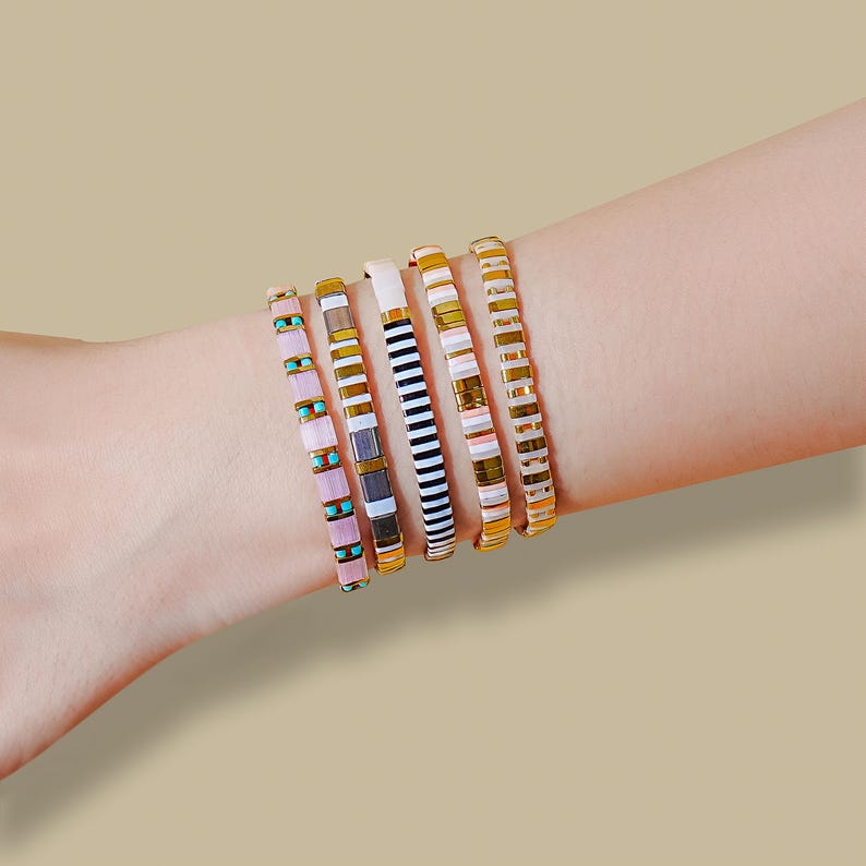 May include: A collection of six colorful bracelets adorns a wrist. The bracelets feature a variety of designs, including stripes, solid colors, and geometric patterns. The color palette includes pink, turquoise, white, black, gold, and brown.
