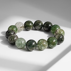 May include: A beaded bracelet featuring round, green gemstone beads. The beads vary in shade, from dark forest green to lighter, translucent green. The bracelet is displayed on a white surface, with shadows adding depth.