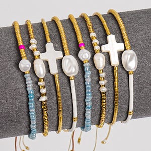May include: A collection of gold beaded bracelets with various charms. The bracelets feature white cross and oval charms, blue and gold beads, and white cord accents. The bracelets are displayed on a gray surface.