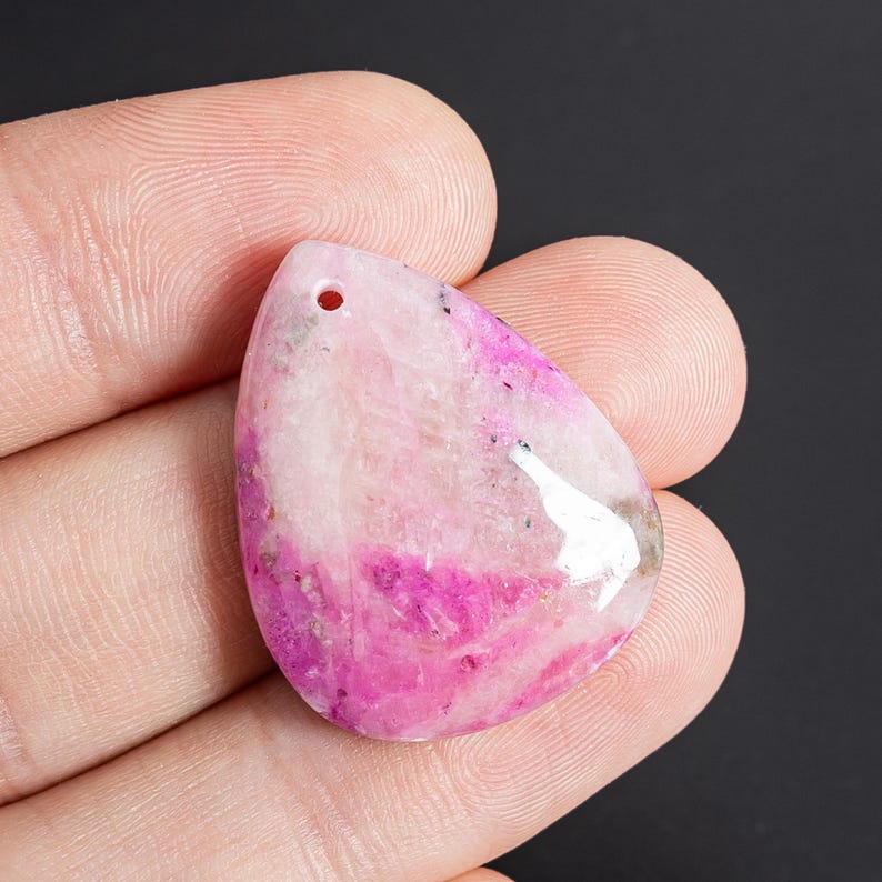 May include: A teardrop-shaped pendant made of polished stone featuring a mix of translucent white and vibrant magenta pink coloration with small dark mineral inclusions and a single drilled hole at the top for suspension or attachment.