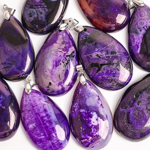 May include: A collection of teardrop-shaped pendants in shades of purple, with silver-colored bails. The polished stones display a marbled pattern with dark purple and black veining. The pendants are arranged on a white surface.