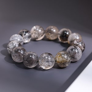 May include: A bracelet featuring clear, round beads with inclusions of black and gold. The beads are strung together, forming a circular shape. The bracelet is displayed on a gray surface.