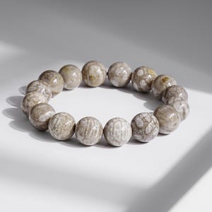 May include: A round bracelet composed of spherical, beige-colored beads. The beads have a marbled appearance with subtle variations in color, ranging from light beige to a darker, earthy tone. The bracelet is displayed on a white surface.