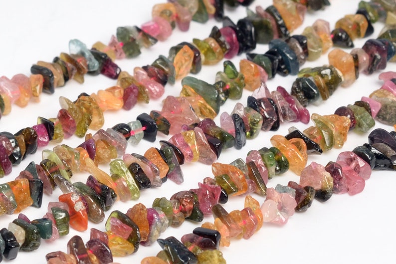 May include: Strands of raw, colorful tourmaline gemstone chips. The chips are a mix of pink, green, orange, and black, strung together. The gemstones are irregularly shaped and have a natural, rough texture.