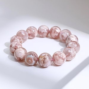 May include: A bracelet featuring large, round beads in shades of pink and white. The beads have a marbled appearance and are strung together on a pink cord. The bracelet is displayed on a white surface.
