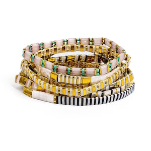 May include: Stack of beaded bracelets in gold, white, black, and pink. The bracelets have a flat, rectangular bead design. Some bracelets have turquoise accents, while others feature a black and white striped pattern.