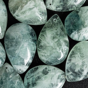 Puede incluir: Una colección de piedras de color verde claro y blanco en forma de lágrima con un agujero perforado en la parte superior. Las piedras son lisas y pulidas, y tienen una apariencia translúcida.