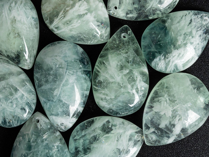 Pu&ograve; includere: Una collezione di pietre di colore verde chiaro e bianco a forma di lacrima con un foro praticato nella parte superiore. Le pietre sono lisce e levigate, e hanno un aspetto traslucido.