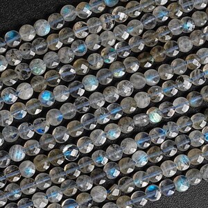 Peut inclure: Une collection de perles rondes facettées en labradorite disposées en rangées parallèles. Chaque perle grise translucide présente une surface polie avec des éclats internes de lumière bleue et dorée irisée, créant un effet scintillant.