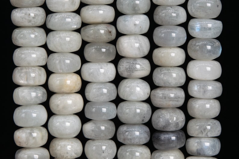 May include: Rows of polished, oval-shaped moonstone beads. The beads are primarily white with subtle gray and translucent areas, creating a soft, luminous appearance. The beads are strung together, ready for jewelry making.