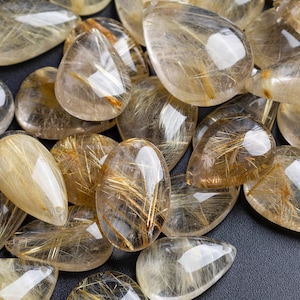 May include: A collection of teardrop-shaped golden rutilated quartz gemstones. The stones are translucent and have a golden shimmer.
