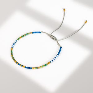 May include: A beaded bracelet with a sliding knot closure. The bracelet features a pattern of blue, white, green, and gold beads. The adjustable cord is silver in color, and the bracelet is displayed on a white surface.