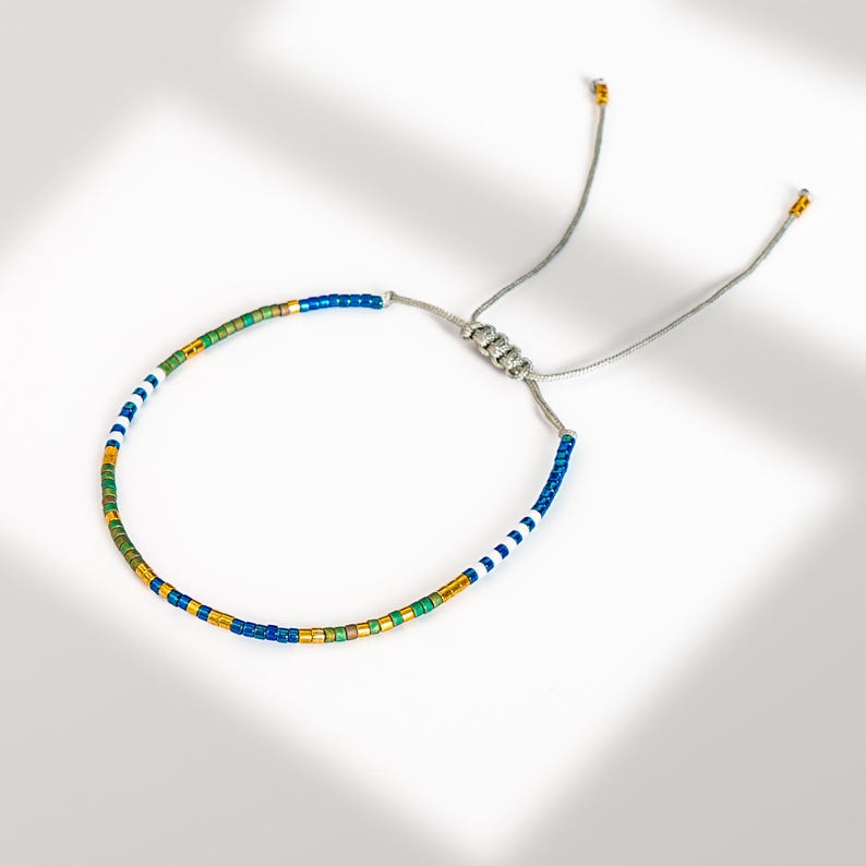 May include: A beaded bracelet with a sliding knot closure. The bracelet features a pattern of blue, white, green, and gold beads. The adjustable cord is silver in color, and the bracelet is displayed on a white surface.