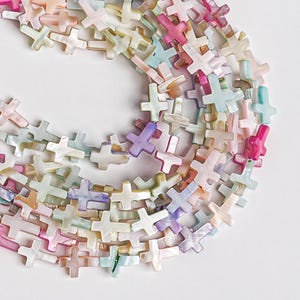 May include: A collection of cross-shaped beads in various pastel colors, including pink, purple, blue, and cream. The beads are made of a pearlescent material, creating a shimmering effect. The beads are strung together, suggesting they are for jewelry making.