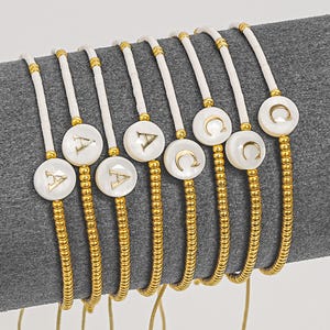 Op de afbeelding: Een verzameling gepersonaliseerde armbanden met witte, ronde, parelmoeren hangers met gouden letters. De armbanden hebben gouden kralenstrengen en een wit koord. De letters zijn "A" en "C".