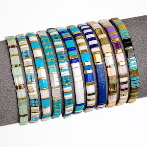 May include: A selection of beaded bracelets in a variety of colours. Each bracelet has a flat, rectangular bead design in shades like turquoise, white, blue, pink, and gold. The bracelets are displayed on a grey surface, highlighting their diverse colour palettes and styles.