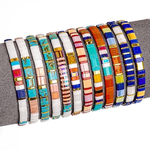 Op de afbeelding: Een verzameling kleurrijke kralenarmbanden. Elke armband heeft een plat, rechthoekig kralenontwerp in verschillende kleuren, waaronder wit, turquoise, roze, goud en blauw. De armbanden worden op een grijs oppervlak gepresenteerd.