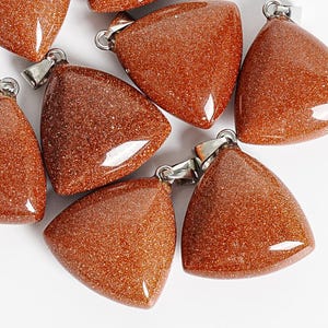 May include: Close-up of several triangular, reddish-brown pendants with a glittery texture. Each pendant has a silver-colored bail attached at the top. The pendants are arranged on a white surface.