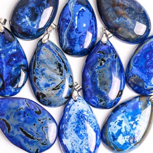 May include: An assortment of blue gemstone pendants with silver-colored bails. The pendants vary in shape and have unique patterns of dark blue, light blue, and gray swirls and marbling. The gemstones are displayed on a white background.
