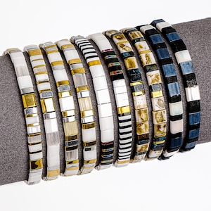 May include: A collection of beaded bracelets in various designs. The bracelets feature square and rectangular beads in white, gold, silver, black, and blue. Some have striped patterns, while others have a mosaic-like appearance. The bracelets are displayed on a gray surface.