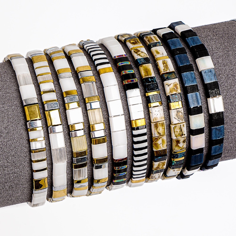 May include: A collection of beaded bracelets in various designs. The bracelets feature square and rectangular beads in white, gold, silver, black, and blue. Some have striped patterns, while others have a mosaic-like appearance. The bracelets are displayed on a gray surface.