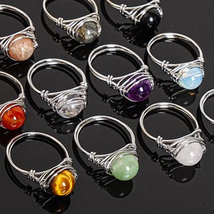 May include: A collection of handcrafted rings with various gemstone beads. Each ring features a silver-colored wire-wrapped design around a different colored stone, including orange, purple, green, and clear. The rings are displayed on a black surface.