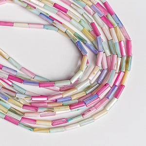 May include: Strands of colorful, iridescent beads in shades of pink, blue, yellow, and green. The beads are cylindrical and arranged in a circular pattern against a white background. These beads are suitable for jewelry making.