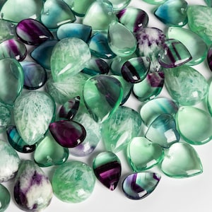 Puede incluir: Una colección de cabujones de piedras preciosas verdes, azules y moradas en forma de lágrima. Las piedras están pulidas y tienen un acabado liso y brillante.