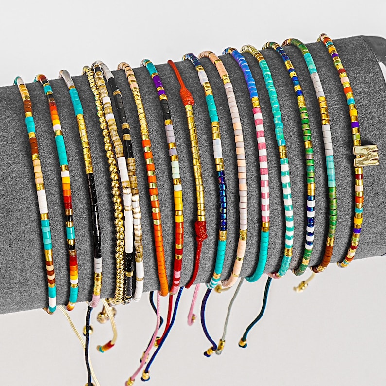 May include: A collection of beaded bracelets in various colors, including turquoise, gold, black, and white. The bracelets are strung on cords and displayed on a gray surface. Each bracelet features a unique pattern of small beads and gold accents.