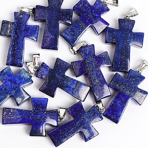 May include: A collection of deep blue lapis lazuli cross pendants with silver-colored bails. The crosses have a polished finish and visible gold flecks. The pendants are scattered across a white surface, showcasing their rich color and texture.