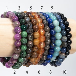 May include: Ten bracelets made of different types of stones, including amethyst, green aventurine, black obsidian, carnelian, tiger's eye, lapis lazuli, aquamarine, blue apatite, and black onyx. The bracelets are stacked on a wooden display stand.