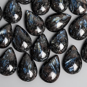 May include: A collection of polished, teardrop-shaped gemstones. The stones are primarily dark with shimmering blue and gold inclusions, creating a striking contrast. The gemstones are arranged closely together, showcasing their unique patterns and textures.