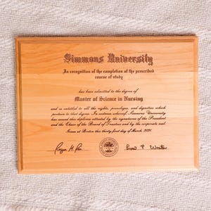 May include: A wooden plaque with the text "Simmons University" and the words "Master of Science in Nursing" engraved. The plaque includes the date "March, 2021" and signatures. The wood has a light brown color.