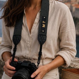 May include: A black camera strap with a textured surface and a rectangular leather patch featuring the gold letters MT. A second camera strap is tan with a brown leather patch engraved with the name Andrea in cursive script.